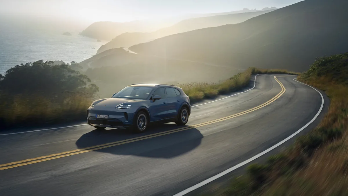 Modrošedý Cayenne S Electric za jízdy po pobřežní silnici v protisvětle, zepředu zleva; vpravo se silnice stáčí do kopce, vlevo pod srázem moře.