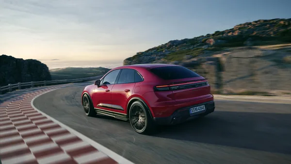 Karmínově červený Macan GTS zezadu zleva při průjezdu zatáčkou; přes celou záď souvislý světelný pruh, vlevo červenobílé obrubníky, v pozadí moře.