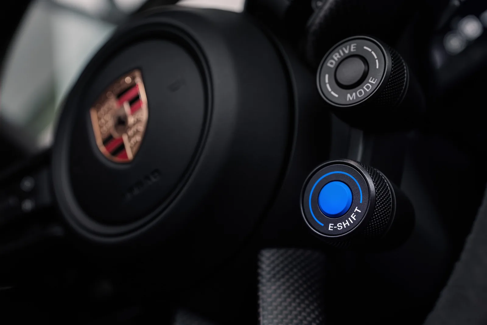 Detail volantu: vlevo znak výrobce ve středu volantu, vpravo dva otočné ovladače — horní s nápisem DRIVE MODE a dolní modře podsvícený s nápisem E-SHIFT.