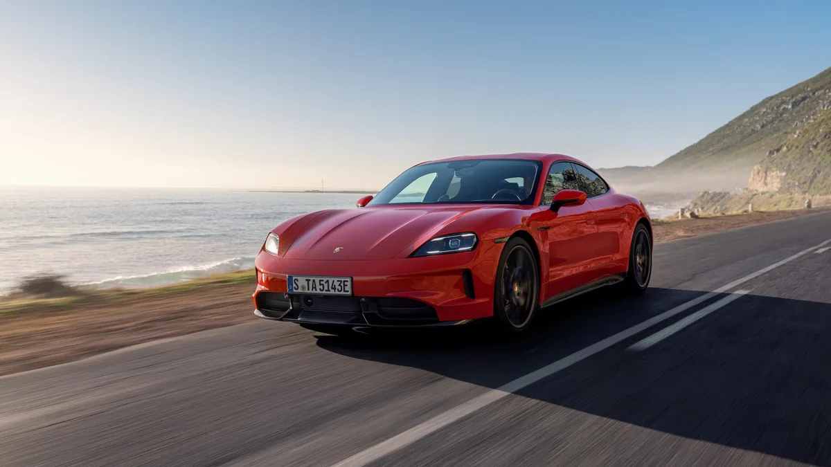Červený Taycan GTS za jízdy po pobřežní silnici; vlevo pod srázem moře, vpravo skalnatý svah, slunce nízko nad hladinou.